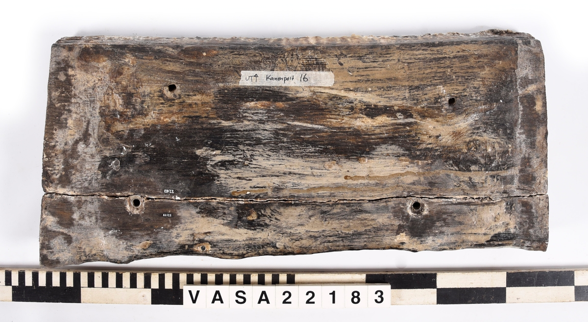 Tröskel till kanonport 16. Paralellogramformad på ändor, fyrkantig på fram och baksidor. 2 klart original spikhål, 6 andra med moderna scruv-spår. 
Splittrad längs en långkant. Kraftig vittrad på alla ytor utomför ändor. Järn impregnering rund alla spikhål på ena yta. Tjock fläckor PEG.