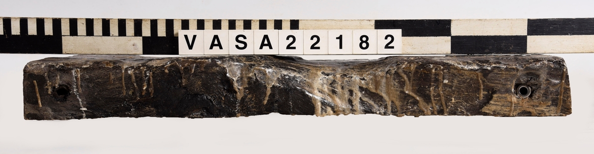 Tröskel till kanonport 14. Nästan rektangular med fasad kanter och ändar. 6 moderna skruvhål, ett klart original spikhål och ett till möjligt.
Lite deformerad, mycket vittrad på ytor (ändar bättre bevarad).