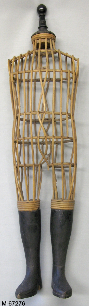 Provdocka av rotting med snidade svartmålade underben/stövlar av snidat trä och svartmålad svarvad avslutning i halsen. Rottingen formad och fastspikad genom vertikala cirklar som håller ut de kroppsformade horisontella rottingbitarna.