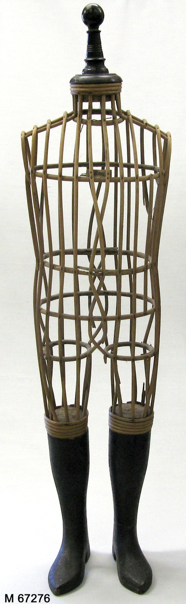 Provdocka av rotting med snidade svartmålade underben/stövlar av snidat trä och svartmålad svarvad avslutning i halsen. Rottingen formad och fastspikad genom vertikala cirklar som håller ut de kroppsformade horisontella rottingbitarna.