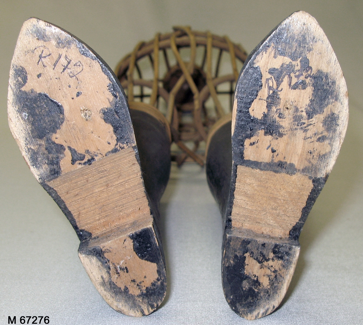 Provdocka av rotting med snidade svartmålade underben/stövlar av snidat trä och svartmålad svarvad avslutning i halsen. Rottingen formad och fastspikad genom vertikala cirklar som håller ut de kroppsformade horisontella rottingbitarna.