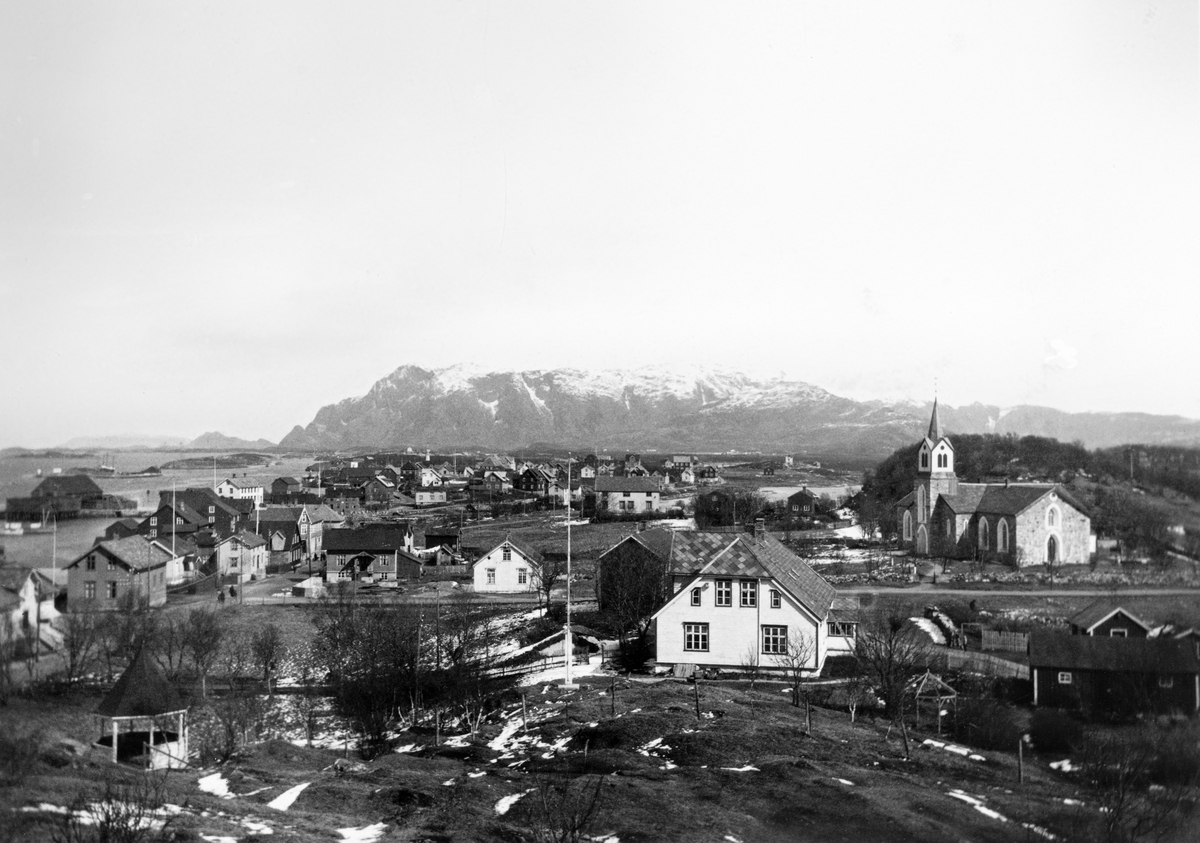Utsikt over tettstedet Brønnøysund, sett fra en bergkolle.  I forgrunnen et stort, kvitt våningshus med halvvalmtak, muligens prestegarden.  Bakenfor til høyre ei lav steinkirke med kvite vindus- og døromramminger, og med tårn på langsida.  Bakenfor og til venstre for våningshuset ligger en del trehus langs ei gate som går parallelt med strandlinja.  I bakgrunnen fjorden og en markant fjellrygg med snø på toppene. 
