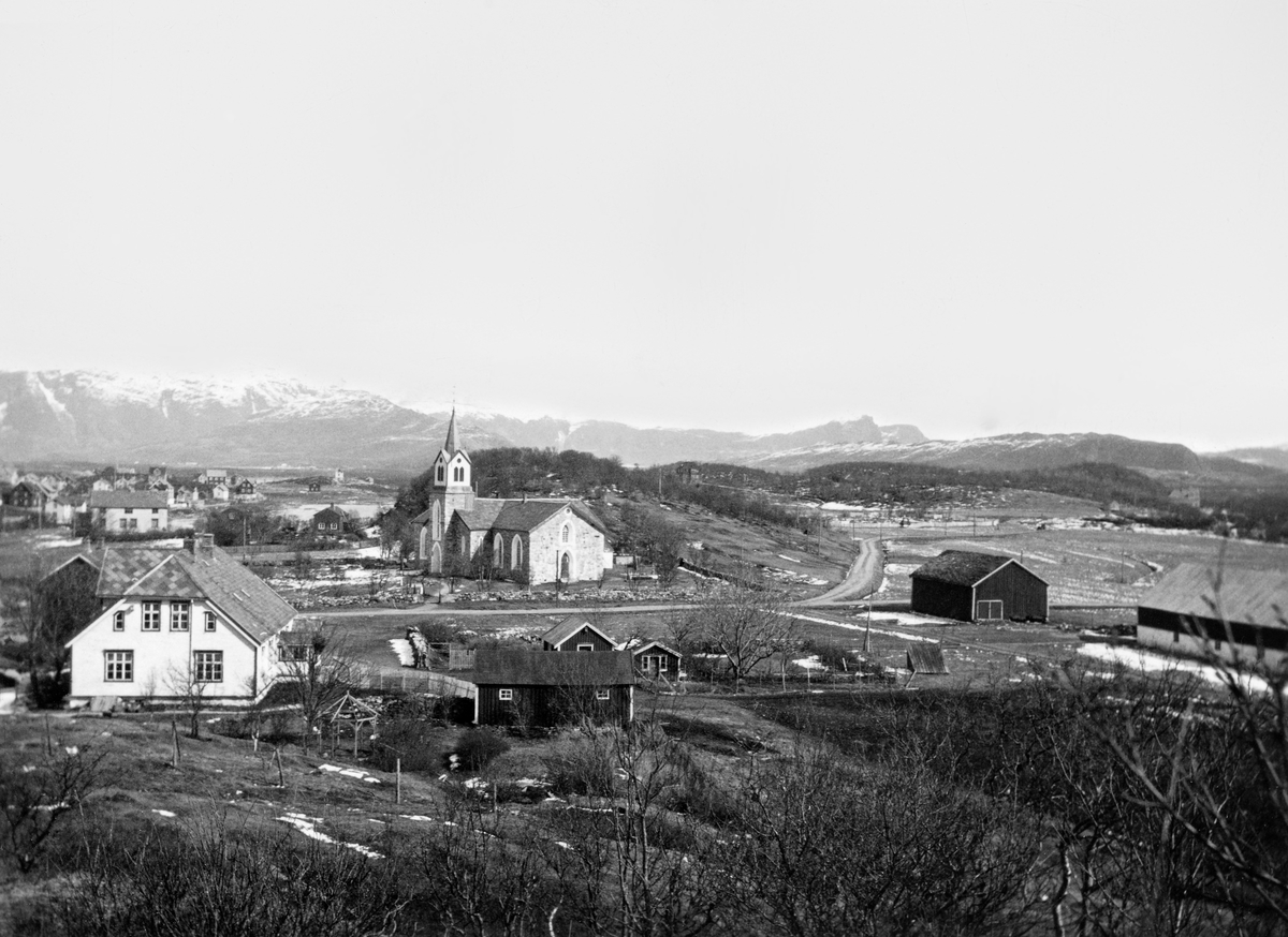 Brønnøy prestegard og kirke.  Det kvitmalte våningshuset på prestegarden ligger til venstre i forgrunnen sammen med en del mindre hus, en større driftsbygning skimtes helt til høyre.  Bakenfor ligger kirka, en forholdsvis lav bygning utført i gråsteinsmur med tårn på langveggen.  I bakgrunnen til venstre skimtes noe av bebyggelsen i Brønnøysund. 