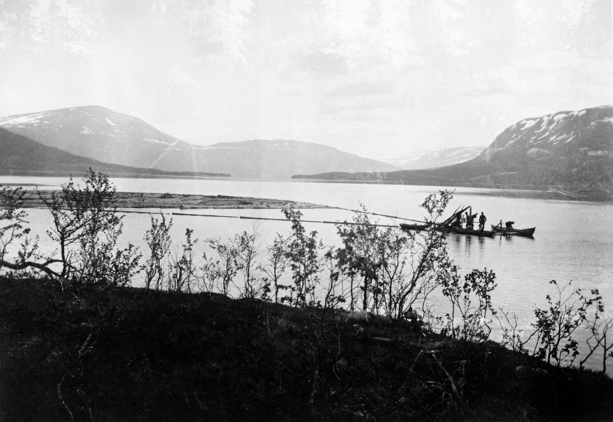 Spillflåtefløting over Unkervatn i Hattfjelldal.  Fotografiet er tatt fra en bakkekam med buskvegetasjon, som dessverre delvis skjuler spillflåten.  På flåten ser det ut til å være tre mann, og en mann står i en båt ved siden av.  Bak flåten ligger en annen robåt.  Der finnes også en tømmerbom med løstømmer som skal trekkes cirka ei mil over vatnet.  I bakgrunnen høye fjell med snøflekker. 