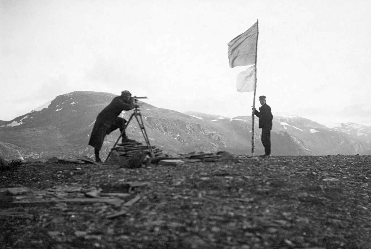 Trianguleringsarbeid i forbindelse med kartlegging av Beiarn statsskog i Nordland.  Fotografiet er tatt på toppen av et høgdedrag.  En frakkekledd mann står bak en oppmålingskikkert, som er montert på et trefotet stativ over ei lita røys.  Noen få meter til høyre for ham står en assistent og holder en bjørkestrange med to flagg på.  Det øverste flagget er av mørkt tøy mens det nederste, som er litt mindre, er av kvitt stoff.  I bakgrunnen ses fjellmassiver der det fortsatt ligger snøstriper i revnene og i skyggefulle lier.  Ifølge påskrift på kartotekkortet datidens Statskog-medarbeidere har arkivert bildet på heter fjellet til venstre Mogdajok, mens det til høyre heter Staupaatind.  Fjelltoppene ble brukt som trianguleringspunkter. Triangulering. 