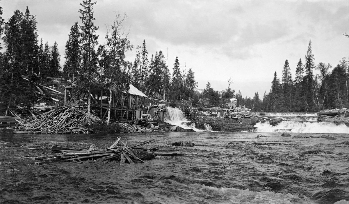 Sagbruk ved Smalåsen i Mellingselva.  Da dette fotografiet ble tatt lå anlegget i Grong kommune i Nord-Trøndelag, i dag ligger lokaliteten i Namsskogan (egen kommune fra 1923).  Det dreier seg om ei vanndrevet sag.  Fotografiet er tatt fra vassdraget, i motstrøms retning.  Saga er plassert i en bindingsverkskonstruksjon med saltak, men uten bordkledning på veggene ved vassdragets venstre bredd.  Bygningen er delvis skult bak to trær.  En del avfallsvirke, antakelig hun, ligger strødd utover bakkeskråningen ved gavlen av saghuset.  Noen hunstubber ligger også ved en stein som stikker opp i elvefaret i forgrunnen.  Noe lengre oppe ses Oterfossen med en ovenforliggende dam.  Ei vannrenne fører fra dammen til sagas vasshjul.  Anlegget ligger i et forholdsvis flatt landskap, bevokst med bjørke- og granskog. 

Reidar Smalås forteller at sagbruket ble reist av Vilhelm Smalås i 1907.  Han fikk en viss pengestøtte fra Grong skogforvaltning.  Anlegget ble komplettert med en høvel omkring 1913.  Den veide cirka sju tonn.  Den gangen var det veg opp til Brekkvasselv og Finvollan.  Derfra var det vegløst, så høvelen måtte transporteres på slede gjennom utmarksarealer og over en strekning på cirka tre mil fram til Oterfossen i Mellingelva.