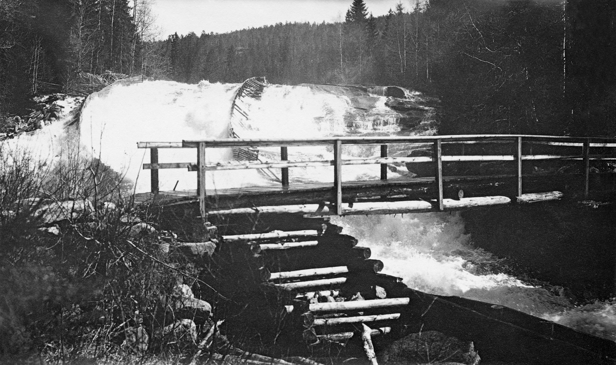 Fra Hogstadfossen i Siljanvassdraget i Telemark.  Fotografiet er tatt i motstrøms retning, mot en foss som faller kvitskummende over en bergrygg.  For å styre fløtingstømmeret gjennom fossen har man bygd tømmerskjermer (skådammer) av tre i den vestre delen av fosseløpet.  Her er det snakk om såkalte "bukkedammer".  I forgrunnen ser vi ei trebru over det ville og flomstore elvefaret.  Brua kviler på ei steinfylt tømmerkister. 