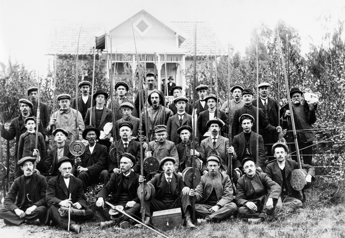 Medlemmer av Hamar Fiskerforening, samlet foran foreningshytta Dalby på Jessnes i Furnes på Hedmarken omkring 1915.  Fotografen har plassert fiskerne på fire rekker slik at de fremste sitter på bakken, de neste sitter i normalhøyde og de bakerste står.  Over og bak de 31 mennene ser vi den nevnte hytta, hvor det også sto et par karer på en veranda.  Fiskerne på bildet er overveiende kledd slik de brukte å være når de fisket, og mange av dem hadde med seg utstyr, spesielt dreggesprøtt med de digre, sjøllagde snellene som ble brukt under slukfiske etter mjøsørret fra båter i langsom, bevegelse.  De fleste av mennene på fotografiet er identifiserte.  Første rekke: Gravør Gustav Gulbrandsen, smed Karl Lukassen, smed Nilsen, A. S. Kristiansen, pølsemaker Ingvald Kristiansen og gullsmed Kolbjørn Berger Christiansen.  Andre rekke: Smed Lenonard Nordrum, Lars Løken, yrkesfisker Anders Furulund, smed J. Hansen, bestyrer Ludvik Monsen, Anders Olsen, kjelsmed Andersen og Alf Bakke. Tredje rekke: smed Lauritz Jørgensen, snekkersvenn Lars Rogstad, Bredor Kristiansen Bjørnstad, Kristian Nilsen, Kr. Hansen, skredder Kristian Kristoffersen, kjemiker Tangen og skiftekonduktør Carl Holter.  Fjerde rekke: mekanikerlærling Martin Knudsen, platearbeider Paul Paulsen, smedsvenn Lars Nordrum, bryggeriarbeider Knut Emilsen, skomakersvenn Bjarne Bakke, ukjent person, Lars Hansen, vognvisitør Halvorsen og kirketjener Andreas Larsen.  Mennene på verandaen skal ha vært Mathias Nilsen og en mann ved navn Larsen.

Den første foreningen for fiskerne i Hamar-området ble stiftet i 1898.  «Hamar og Omegns Fiskeriforening» skulle først og fremst organisere motstanden for et grunneierinitiert forbud mot oterfiske og dregging i flere av kommunene på østsida av Mjøsa (1886).  Fiskerne hadde ingen respekt for forbudet, og et strengt oppsynsregime gjorde at mange av dem ble bøtelagte.  Hamar og Omegns Fiskeriforening sørget for juridisk bistand til de bøtelagte fra advokaten Johan Zeier, noe som førte oppsynet over på defensiven. Dermed ebbet virksomheten i foreningen ut.  Den ble imidlertid reorganisert i 1907 med den driftige fiskeentusiasten Ludvik Monsen (i andre rekke på dette fotografiet) som leder og under navnet «Hamar Fiskerforening». Året etter ble dreggeforbudet opphevet.  I 1910 kjøpte foreningen hytteeiendommen Dalby på Jessnes av et av medlemmene, Carl Knudsen (lengst til høyre i tredje rette på dette fotografiet).  Dalby hadde imidlertid ingen strandrett, så det ble også bygd ei lita hytte på Nes-sida av Furnesfjorden, før man i 1919-1920 kjøpte eiendommen Fagervik, like ved Dalby, hvor Hamar Fiskerforening bygde det som kom til å bli et mer varig samlingssted for medlemmene.