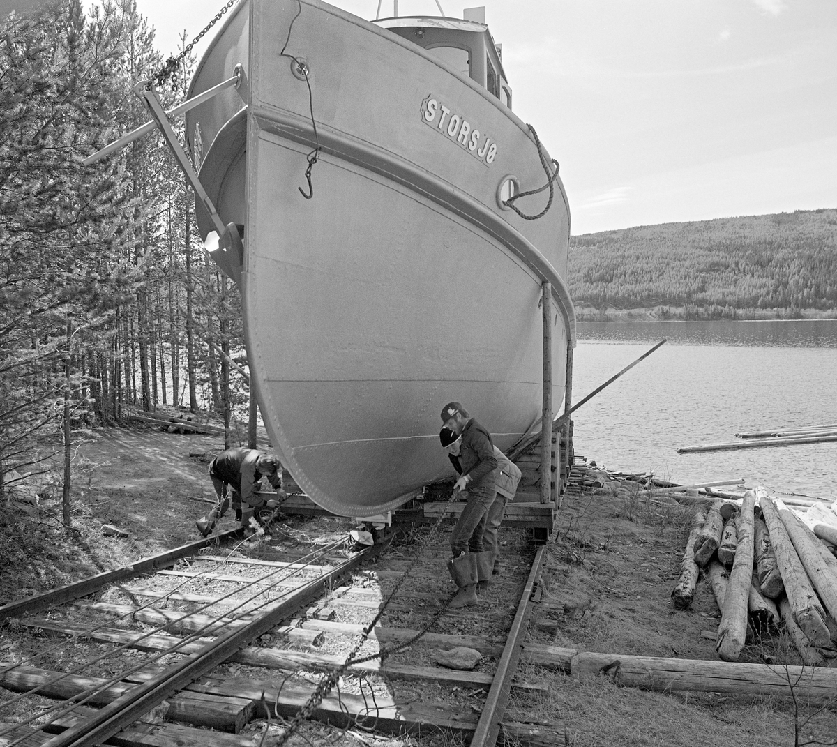 Sjøsetting av tømmerslepebåten «Storsjø» på innsjøen den var oppkalt etter i Rendalen i Hedmark.  Fotografiet er tatt våren 1984, som var den siste fløtingssesongen i denne delen av Glommavassdraget.  I vintersesongen lå båten i opplag på en slipp ved den såkalte «Dampbåtvika» på østsida av Storsjøens nordende.  Her var det lagt en skinnegang på tresviller i en slak skråning ned mot sjøen og et stykke videre under vannflata (til venstre i dette bildet).  På denne skinnegangen gikk det ei vogn med jernbanehjul.  Vogna var forankret i en kraftig vaier med forbindelse til en vinsj et stykke oppe på land.  Ved hjelp av denne vinsjen kunne vogna, med båten som last, kjøres kontrollert ned mot sjøen eller opp igjen på tørt land.  Til høyre i forgrunnen lå det en del værslitt lensetømmer.  Direksjonen (styret) for Christiania Tømmerdirektion (Glomma fellesfløtingsforening) vedtok i 1911 å gå til anskaffelse av en dampslepebåt som kunne brukes til å trekke tømmer i bommer over den lange og djupe Storsjøen. Her hadde det vært vanskelig å få fløtt fram virket i løpet av en sesong, noe som innebar verdiforringelse av tømmeret. Det nye fartøyet ble kontrahert fra Glommens mek. Verksted i Fredrikstad. I mangel av farbar vannveg mellom verftet og sjøen der båten skulle brukes ble komponentene fraktet med tog til Koppang. Derfra ble de trukket på sleder over til Burua i Ytre Rendalen, hvor komponentene ble klinket sammen. D/S Storsjø ble 58 fot 6 tommer lang og 12 fot bred.  Ved normal belastning gikk kjølpartiet 6 tommer under vannskorpa.  Båten ble satt i drift våren 1912 og gikk som tømmersleper i Rendalen fram til 1984.  Mer informasjon om denne slepebåten og det transportsystemet den inngikk i finnes under fanen «Opplysninger».