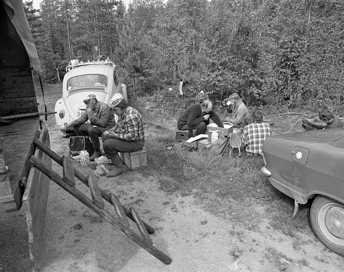 Arbeidslaget som fløtet tømmer i elva Søndre Osa i Åmot i Østerdalen i 1981, fotografert under ei mat- og kvilepause ved Kvernfallet. Glomma fellesfløtingsforening hadde organisert dette slik at Paul Solberg fulgte fløterne med en liten lastebil, der han hadde ryggsekker med mat, kasser til å sitte på og eb del annet utstyr. Paul stanset bilen på steder der det var høvelig å tenne kaffebål og slappe av litt når karene tok seg tid til pauser. Her ser vi lastebilen med en liten stige opp mot lasteplanet til venstre i forgrunnen. Nærmest bilen satt Arne og Arnstein Norsted, far og sønn fra Osen, på hver sin trekasse, der de spiste brødskiver og drakk kaffe eller mjølk. Litt lengre til høyre ser vi Nikolai Myrene, også han fra Osen, med handa oppi kjølebagen sin. De andre tre som satt eller lå i graset framfor Myrene var unge karerer fra Flisa-området i Solør, som hadde tatt turen nordover for å hjelpe til med fløtingsarbeidet: Kenth Mokkelbost, Dag Tore Syversen og Tor Anders Syversen. Karene var arbeidskledde. En av dem hadde vrengt av seg støvlene for å kunne tørke strømpene på ei lita furu i bakgrunnen. Folkevognbobla tilhørte sannsynligvis museets fotograf, OT Ljøstad.

I 1981 ble det levert 10 260 kubikkmeter med tømmer - slipvirke til treforedlingsbedriften Borregaard i Sarpsborg - ved Osensjøen. Dette tømmeret skulle fløtes ned den cirka 17 kilometer lange Søndre Osa, over i den nedre delen av Renaelva, som skulle føre det ut i hovedvassdraget, Glomma. Arbeidslaget som påtok seg denne oppgaven besto dels av lokale folk fra Osen, dels av unge, entusiastiske fløtere fra Flisa-traktene i Solør.