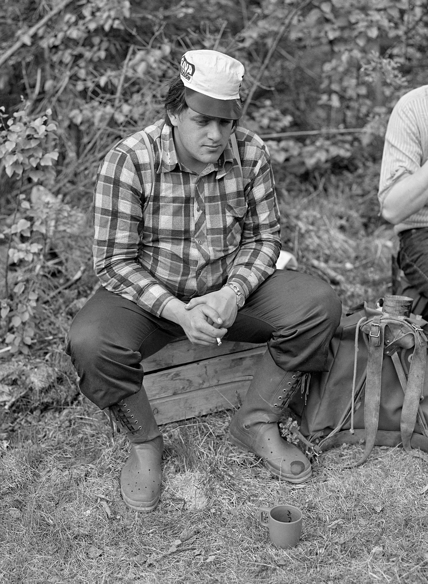 Arnstein Norsted (1958-2022) fra Nordre Osen, fotografert i ei kvilepause under tømmerfløtinga i elva Søndre Osa i 1981. Norsted satt på ei trekasse. Han var kledd i beavernylonbukser og rutete flanellskjorte. På hodet hadde han ei arbeidslue med en plastskjerm som skulle skjerme for solstrålene. Lua var en reklameartikkel fra selskapet Glava, som produserte bygningsisolasjon. Fottøyet var gummistøvler, som ifølge fotografen hadde hull slik at vann som trengte inn i dem når fløterne måtte ut på steder der det var djupt også skulle dreneres ut igjen. I pausa slappet Norsted av med en sigarett.