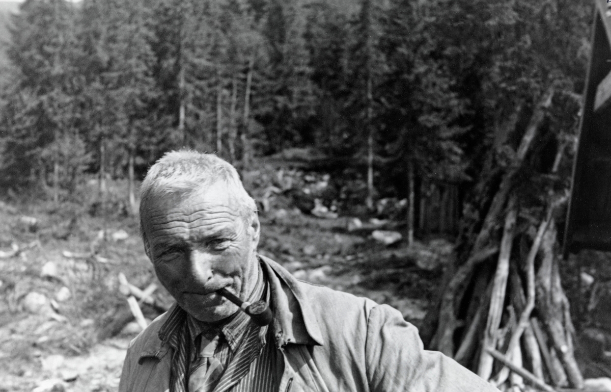 Damfløter Simen Nyutstumoen (1880-1953) fotografert ved Finstaddammen i Øvre Rendalen.  Da dette bildet ble tatt var Simen cirka 60 år gammel. Han hadde kortklipt, grått hår og pipe i munnen.  Han var iført ei slitt arbeidsjakke.  Under jakka bar han busserull, skjorte og slips.  Helt til høyre i bildet skimtes takutskottet i gavlen på damkoia og et reis med grånet ved.  I bakgrunnen granskog.  Simen Nyutstumoen var fra Tynset.