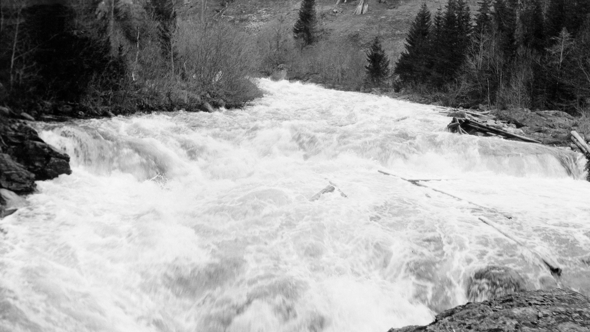 Finnstadfallet i Finnstadåa i Øvre Rendalen.  Fotografiet viser et flomstort elveløp med kvitskummende overflate.  I forgrunnen passerer vannet en terskel i eleveleiet som danner et lite (kanskje meterhøyt) fall. Et par tømmerstokker skimtes i det skummende vannet nedunder fallet.  Opptak med nummer SJF. 1990-00125, SJF. 1990-00126 og SJF. 1990-00141 er gjort på samme sted. 