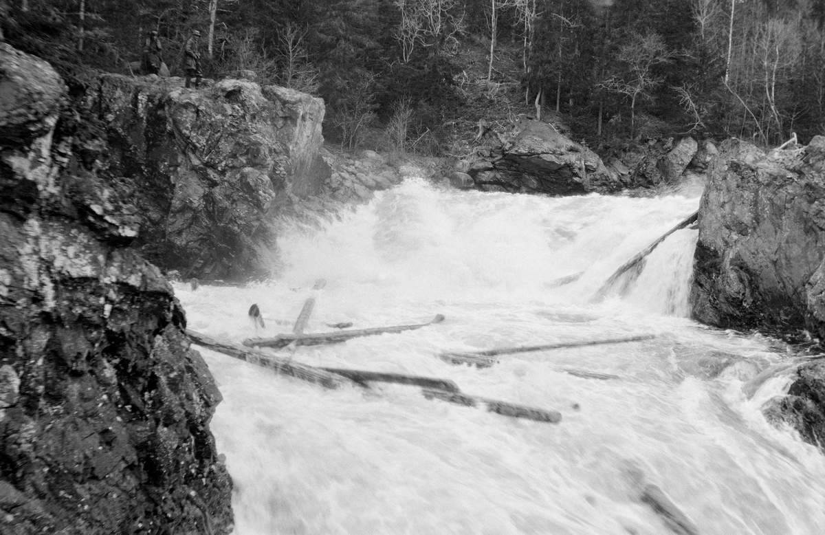 Fra Finnstadfallet i Finnstadåa i Øvre Rendalen.  Fotografiet viser et lite fossefall på et sted der elva går flomstor mellom to bergnabber.  Også nedenfor fallet er elva preget av et strømkav.  Her ses en del tømmerstokker som flyter mot neste terskel i elveløpet (nederst i bildeflatas høyre hjørne). På bergnabben til venstre for fallet står ei lita menneskegruppe og betrakter flommen og fløtingstømmeret.  Elveleiet er ellers omgitt av granskog ispedd enkelte bladlause lauvtrær.  Opptaket er gjort på samme sted som SJF. 1990-00139.  Jfr. også SJF. 1990-00123 og SJF. 1990-00124. 