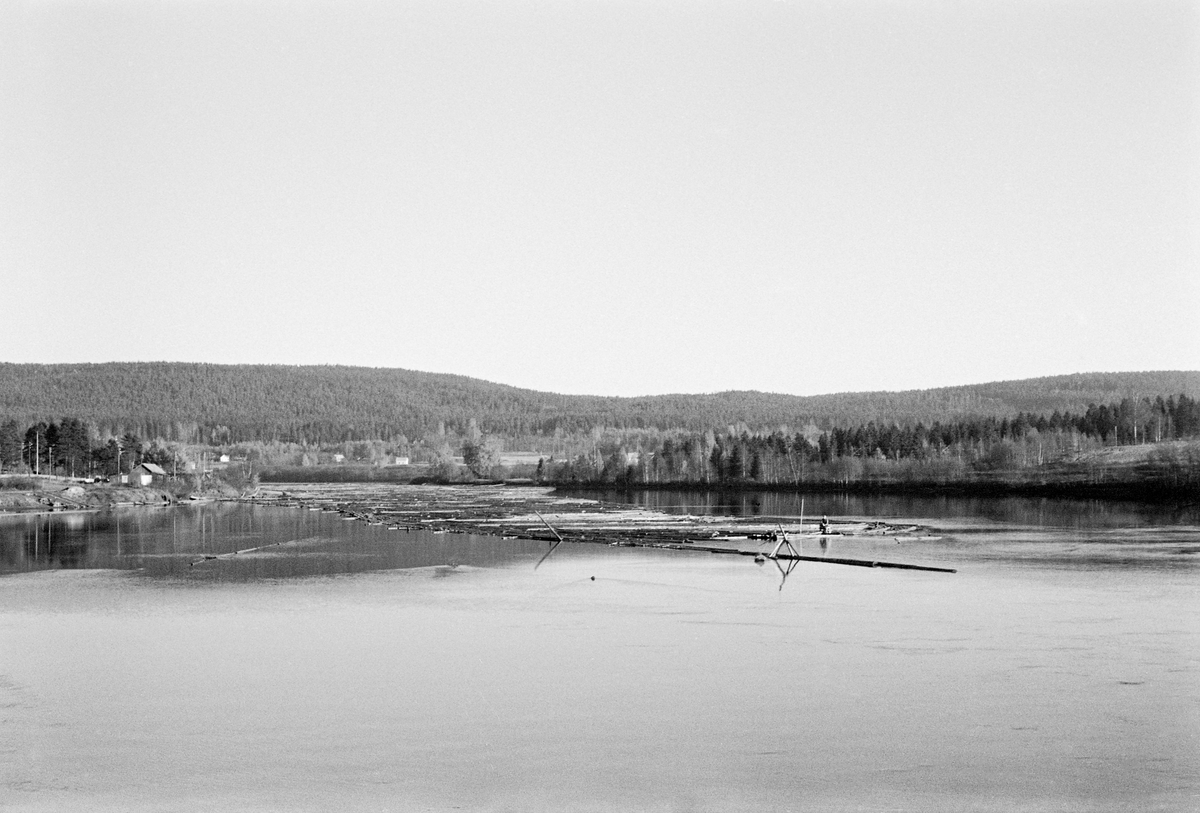 Fra lenseanlegget ved Funnefossdammen i Glomma i Nes kommune på Romerike.  Fotografiet ble tatt i 1959.  Det viser et stilleflytende, blankt vannspeil spm ble brutt av et lensanlegg som holdt en del tømmer i en posisjon som skulle være optimal for videre fløtig gjennom den nedenforliggende fossen.  I bakgrunnen ser vi en del hus og åkerarealer langs vassdraget og skogkledde åser bakenfor.