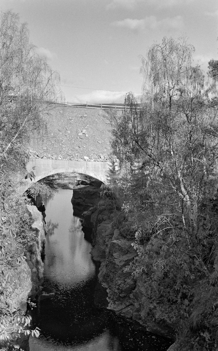 Ny vegbru for Rustvegen (fylkesveg 256) i Nord-Fron kommune i Oppland, der denne vegen krysser elva Vinstra ved Loholet, fotografert høsten 1959. Dette er en lokalitet der elva renner gjennom et trangt skar med steile bergskrenter på begge sider.  Dette var for så vidt et takknemlig sted å bygge bru, både fordi elva var smal, slik at bruspennet kunne gjøres kort, og fordi det var meget stabil byggegrunn.  I dette tilfellet valgte man å bygge ei betongbru med oppoverbuet underside.  På den plane oversida ble det lagt ei ganske tjukk grusbane for vegen.  Da dette fotografiet ble tatt var autovernet begrenset til et spinkelt tregjerde.  Loholet hadde i alle år vært et problematisk sted for tømmerfløtinga, fordi stokkene hadde lett for å sette seg fast i den trange passasjen, hvor det på grunn av de steile elvebreddene var vanskelig og risikofylt å løsne dem.  Når det gjaldt brua var det nok essensielt for Glomma fellesfløtingsforening at den fikk såpass høyde at den ikke kom til å bli til sjenanse for fløtingsarbeidet, noe man ser ut til å ha oppnådd.