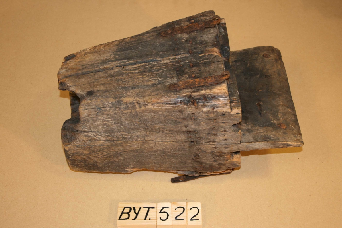 Firkanta trestykke med røyrforma hol gjennom. Mange spikrar, restar av beslag. Sprekt, delvis roten, brannskader ytst.