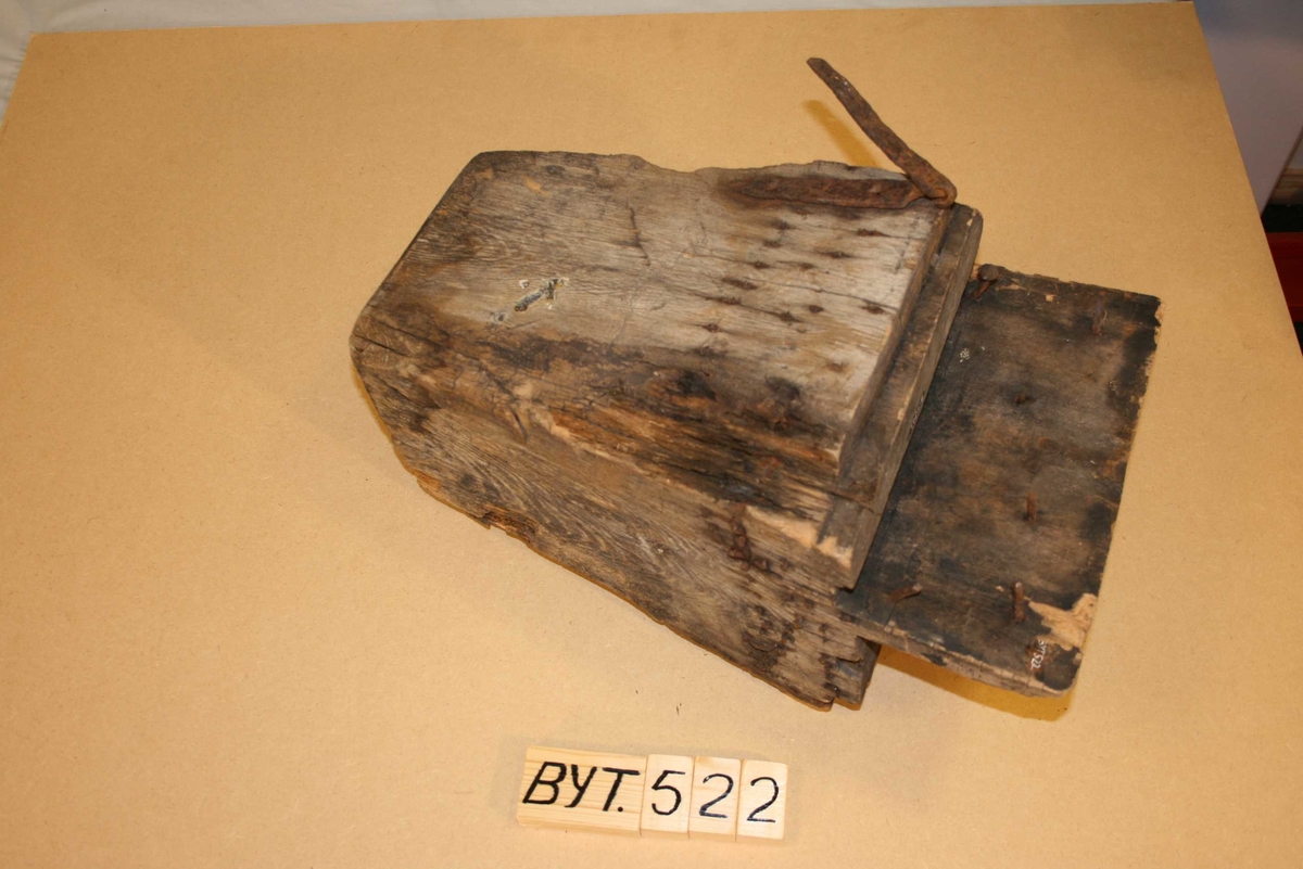 Firkanta trestykke med røyrforma hol gjennom. Mange spikrar, restar av beslag. Sprekt, delvis roten, brannskader ytst.