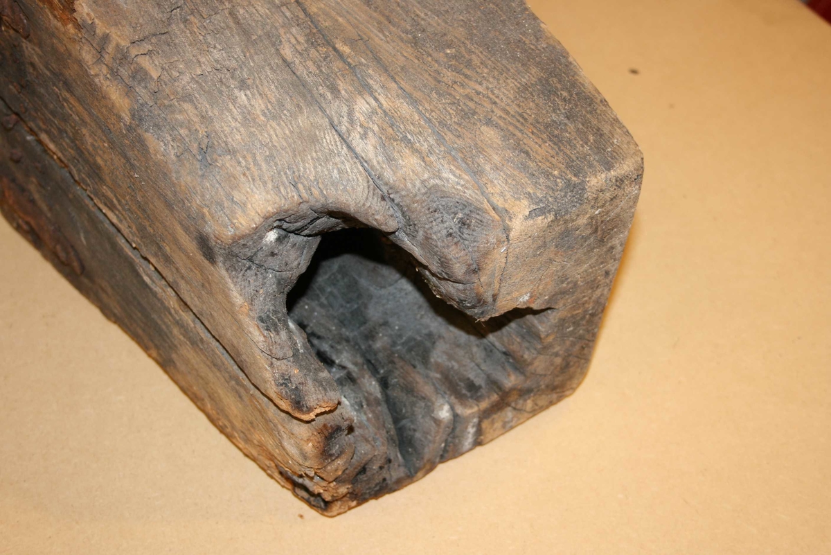 Firkanta trestykke med røyrforma hol gjennom. Mange spikrar, restar av beslag. Sprekt, delvis roten, brannskader ytst.