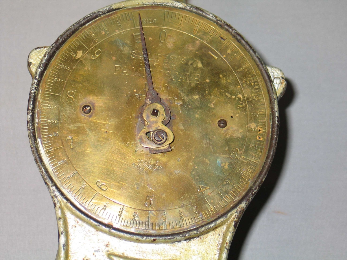 Rund fotplate, metall. Klokkeforma talskive med vektskala i gram og kilo, med visar. Sjølve talskiva av messing. Skål på toppen. Grågrøn måling.