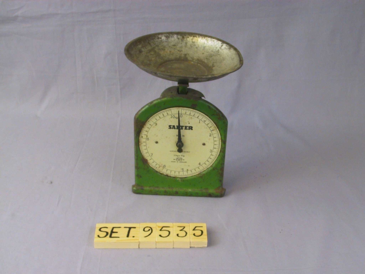 Flat framside med rett botn og rette sider, avrunda topp. Kvit, klokkeforma talskive med vektskala og visar. Laus skål som står på kryssforma, flate berepinnar på toppen av stav som går ned i halvsylinderforma bakstykke. Grønmåla med kvit talskive og svarte tal.