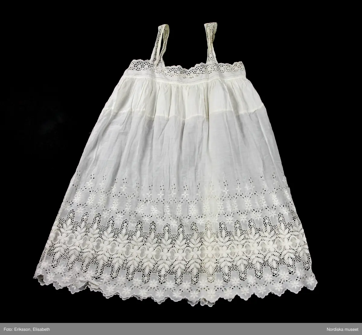 Underklänning av bomull och med engelskt broderi. Rak klänning med två smala axelband (ca. 4 cm bredd), också med engelskt broderi i ett blommigt mönster. Kring urringningen samma mönster, liksom en bård nedtill på klänningen. Rak underklänning som följer rådande stilideal under 1920-talet.
/Petrine Knight & Helena Lindroth, 2014-02-10