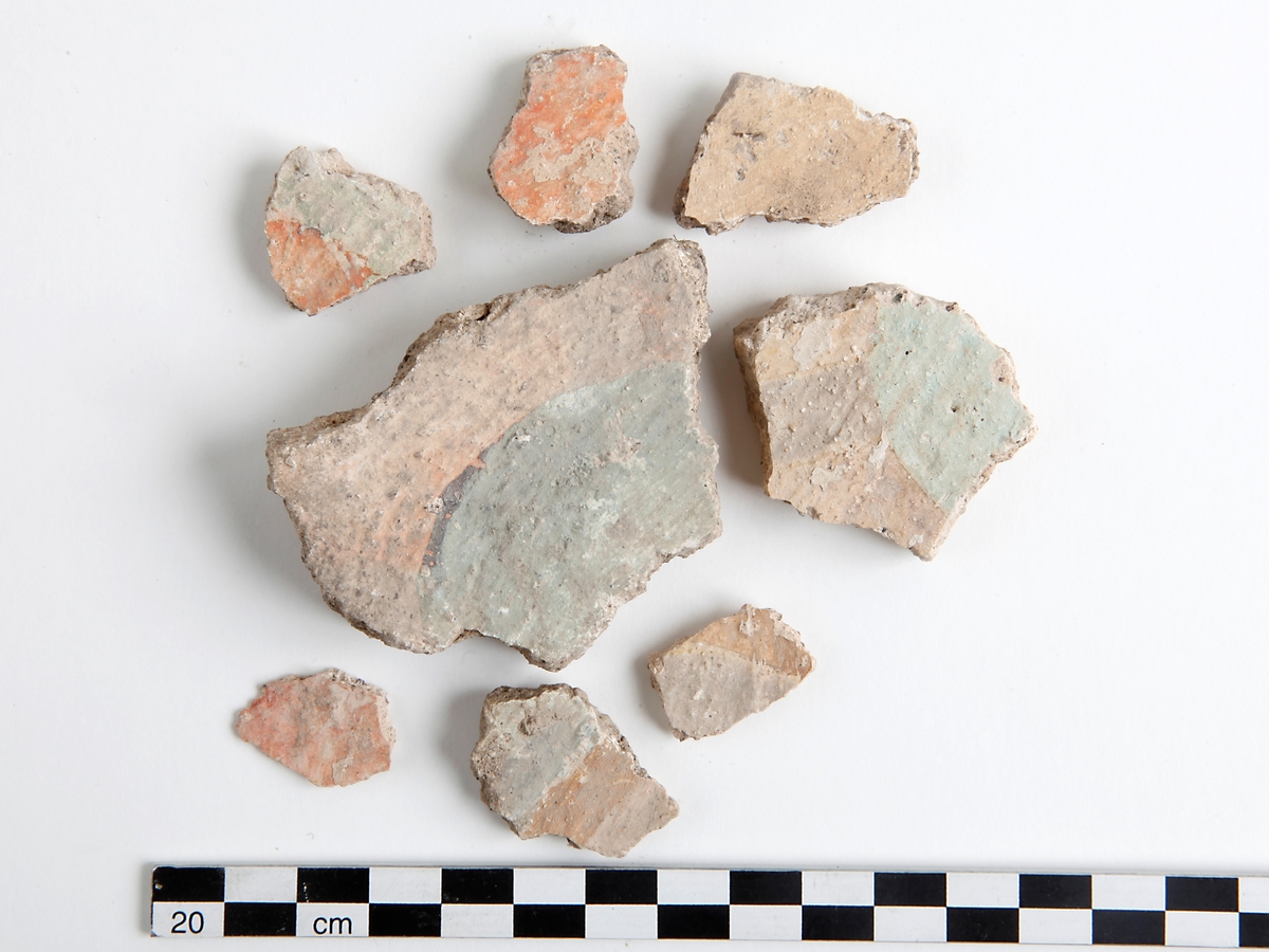 Fragment av kalkputs. Bemålade kalkputsfragment med schablonmotiv samt heltäckande färger i rött, grönt/turkos, gult, orange, svart, och brunt. Datering sent 1400-tal.