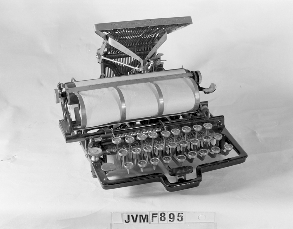 Skrivmaskin av amerikanen Fitchs patent, där typerna är placerade ovanför och bak maskinen och slår mot valsen uppifrån. Maskinen är av stål och svartlackad metall, med detaljer i guld. Valsen är klädd med svart gummi. Ett gulnat papper sitter kvar kring valsen.

Modell/Fabrikat/typ: 3057