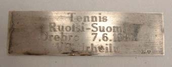 Bordsstandar med blå treflikig flagga. I mitten tryck av en vit bevingad oval. Vita tofsar nertill. Sockel av grå marmor med en silverplatta med följande text på finska:
"Tennis
Ruotsi-Suomi
Örebro  7.6.1980
VR Urheilu".

Ungefärlig översättning: "Tennis Sverige-Finland Örebro 7.6.1980 VR idrott".