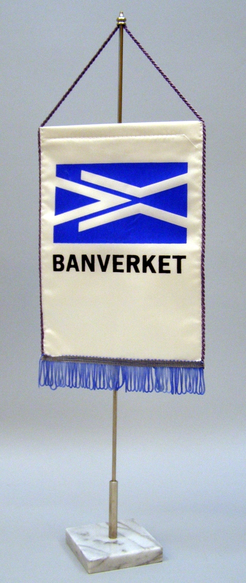 Borsstandar med rektangulär flagga av siden med Banverkets logotyp som är blå mot vit botten och med texten "BANVERKET" nedanför. Nedtill en ljusblå frans, på långsidorna en lila snodd som är fäst upptill på vertikal stång av metall. Stången har en kvadratisk fot av ljus gråspräcklig marmor. Går att montera isär.