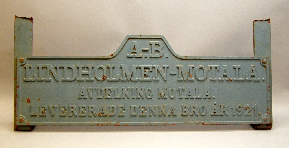 Skylt i järn målad i blå-turkose nyans, rektangulär (förutom en upphöjning i mitten ovantill), med upphöjd kant längs med ytterkanten samt text i relief: "A. - B. LINDHOLMEN - MOTALA. AVDELNING MOTALA. LEVERERADE DENNA BRO ÅR 1921." På baksidan avkapade fästen i järn målade i samma nyans som skylten.

Skylten har varit monterad på klaffbro över Södertälje kanal som rivits år 2010 i samband med att ny bro byggts och tagits i bruk. Vid rivningen av bron togs skylten tillvara då man tyckte det var synd att den kasserades. Skylten satt monterad på tvärbalken mot kanalen vid motviktsstödet. Den monterades ned genom att fästena kapades. Den förvarades sedan en kort tid på byggarbetsplatsen i Södertälje innan den togs om hand av Sveriges Järnvägsmuseum. Klaffbron konstruerades och levererades av A-B Lindholmen - Motala, avdelning Motala och togs i bruk 1921, Västra stambanan (Statens Järnvägar, bandel 336). Enligt Trafikverkets hemsida har denna bro krävt mycket underhåll, vilket har stört järnvägstrafiken. Bron uppfyllde inte heller de krav som idag ställs för järnväg och sjöfart, den klarade inte alltför tunga tåg och större djupgående fartyg hade svårt att passera. En ny bro började byggas våren 2007 av Banverket och den var klar år 2010 då den gamla bron revs av Trafikverket (Trafikverket bildades våren år 2010 och innefattar bl. a. verksamheten vid tidigare Banverket).
Källor:
Webben: www.trafikverket.se "Södertälje kanal, ny järnvägsbro", Informationen hämtad 2010-11-22
Litteratur: Järnvägsdata med trafikplatser, Svenska järnvägsklubben nr 83, 2009, s330
Litteratur: Våra Broar - en kulturskatt, 2001, s394-397.
Litteratur: Statens Järnvägar 1906-1931, del 2 Bana och banbyggnader, s176-177.
Webben: www.wikipedia.se "Södertälje kanal", informationen hämtad 2010-12-01

I Sveriges Järnvägsmuseums samling finns en modell av den klaffbro som skylten har suttit på, med inventarienummer Jvm 9286.