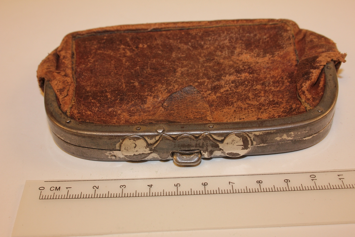 Rektangulær/oval lommebok i skinn. Hengsla metallbøyle med knapplås. Innvendig hengsla metallbøyle som har delt lommeboka i to rom. Skinnet som har fungert som skillevegg, festa til innvendig hengsla bøyle, er borte. Skinnklaff ved opning (for å lette opning av lommebok).