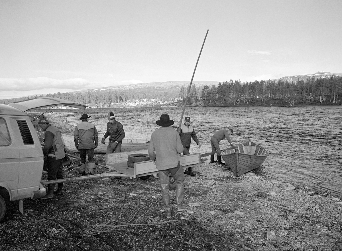 Tømmerfløtere ved Femundselva i Trysilvassdraget våren 1986.  Fotografiet er tatt på velteplassen ved Røskjota i Engerdal, dit karene hadde kjørt med bil for å kunne starte med sluttrensken.  Da dette bildet ble tatt var fløterne i ferd med å sette en fløterbåt på vannet.  Den hadde de kjørt med seg på en tilhenger bak en Volkswagen Transporter.  Båttypen var om lag fem og en halv meter lang, bygd av forholdsvis smale, tynne furubord som var sammenføyd ved hjelp av «kinger» (jernkramper) i steden for klinknagler.  Det var fløterbasen Kåre Joar Graff som bygde slike båter, et handverk han hadde lært av onkelen Ole K. Rømoen.  I 1986 var det 23 mann som deltok i fløtinga på Femunds- og Trysilelva.  På sidevassdragene var det ikke lenger fløting.  Da dette fotografiet ble tatt var karene klare for sluttrensken, som innebar at stokker som hadde strandet langs elvebreddene eller på stein- og grusører ute i elveløpet skulle løsnes, slik at de kunne flyte med strømmen ned mot Karlstad.  Båtene var gode å ha når fløterne skulle nå tømmer som satt fast midt ute i elveløpet eller på motsatt side av vassdraget i forhold til der hvor de befant seg.  Hver båtlag besto av fire mann: en «framstavnkar» ved årene, en «bakstavnkar» med skåtestang og to «haugkarer» med fløterhaker.  I tillegg til båtlagene gikk 4-5 karer langs hver elvebredd og løsnet stokker som hadde satt seg fast der.  På den terminalplassen der dette fotografiet er tatt lå det et par uker tidligere cirka 15 000 kubikkmeter tømmer som ble utislått ved hjelp av hjullastere (jfr. f. eks. SJF.1994-00019).