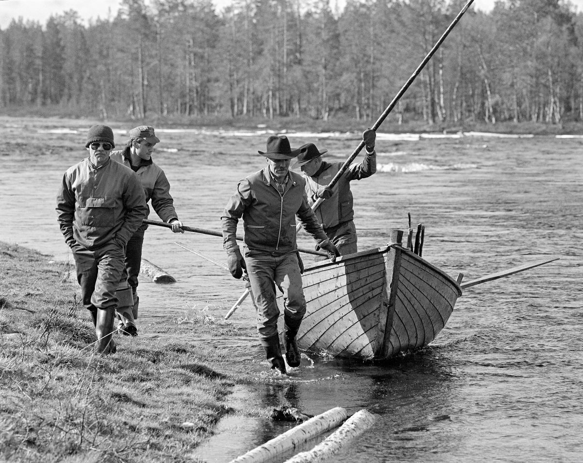 Båtlag ved Myrstadvelta i Femundselva våren 1986. Bildet viser fire menn, hvorav den ene sto i båten, mens gikk i strandsonen.  Kåre Joar Graff dro tilsynelatende båten i motstrøms retning i det stilleflytende elvevannet.  Båtlagene i Trysilvassdraget besto av fire mann – en «framstavnkar» ved årene, en «bakstavnkar» som bisto med manøvrering ved hjelp av ei skåtestang fra akterenden og to «haugkarer», som etter hvert skulle over på tømmerhauger som hadde strandet i den sentrale delen av elveleiet for å arbeide stokkene tilbake til de frittstrømmende vannmassene for å løsne stokkene.  I den perioden da dette fotografiet ble tatt var det arbeidsformann Kåre Joar Graff som bygde fløterbåtene som ble brukt i dette området, slik han hadde lært det av onkelen Ole K. Rømoen, som også var fløterveteran.  Båttypen var forholdsvis bred og lang, men likevel lett.  Båten var lagd av forholdsvis smale og tynne furubord, som var sammenføyd ved hjelp av «kinger» (metallkramper).