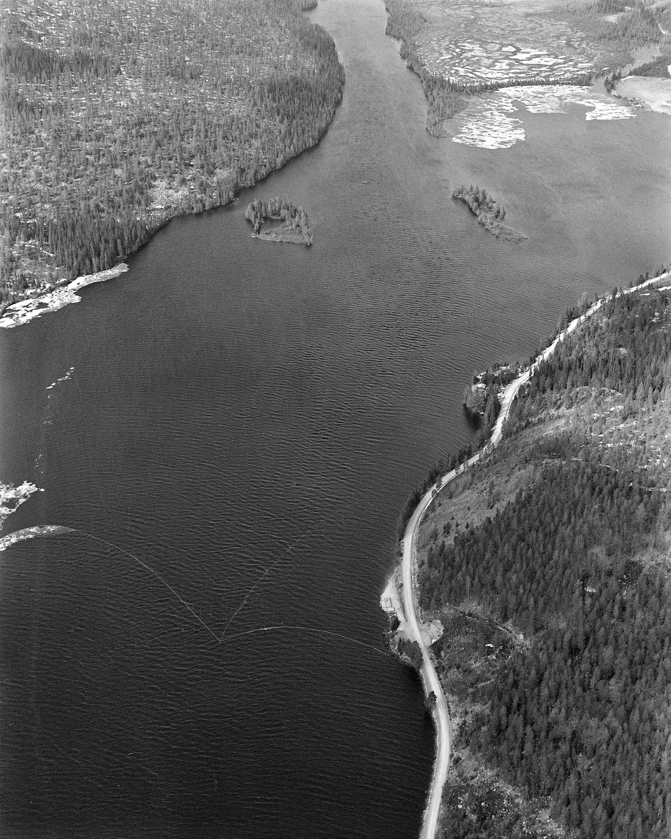 Flyfotografi, tatt over stopplensa i nordenden av Sennsjøen, der fløtingstømmer fra Engerdal ble holdt tilbake i påvente av at forholdene lå til rette for fløting videre sørover Trysilelva. Bildet er tatt i midten av mai 1986, på et tidspunkt da det fortsatt lå litt is langsmed den ene sida av sjøen, men før tømmeret fra ovenforliggende skogområder hadde nådd ned hit.  Til høyre i forgrunnen (på østsida av Sennsjøen) ser vi litt av fylkesveg 581 (mellom Sundet i Engerdal og Engerneset i Trysil).  Holmene nord for lensa kalles Sandholmene.

Sennsjølensa samlet opp alt tømmeret som kom på Femundselva fra Engerdal.  Der var man avhengige av å fløte mens det var flomvannføring, for å slippe at tømmeret satte seg fast på grusører i elveløpet.  Lengre nedover var det et poeng at vårflommen skulle være på retur før de store tømmermengdene kom.  I det flate lendet langs Trysilelva kunne nemlig flomvannet føre mange av stokkene langt inn på land, og i så fall ble det arbeidsomt for fløterne å bære dem tilbake til vannstrømmen i elveløpet når vannstanden sank.  Derfor ble engerdalstømmeret ofte holdte tilbake i Sennsjølensa til begynnelsen av juni.  Det året dette fotografiet ble tatt ble det levert om lag 22 000 kubikkmeter fløtingsvirke fra Engerdal.  Dermed ble bilvegene i regionen spart for 3 - 4 000 lastebillass.  Fløterne var avhengige av nordavind når det åpnet Sennsjølensa, for strømdraget var ikke kraftig nok til å dra tømmeret gjennom sjøen om det blåste fra sør.  Dette fotografiet ble etter at lensa var lagt, men før tømmeret nordfra hadde nådd Sennsjøen.