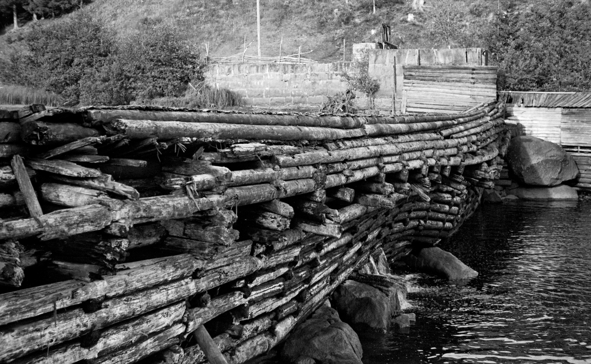 Syversætredammen i elva Flisa i Åsnes i Solør, fotografert fra nordvestre elvebredd i 1955.  Bildet ble tatt i en tørkeperiode midt på sommeren, da vannføringa i elva var minimal, slik at damkonstruksjonen var godt synlig, uten overløpende vann.  Dermed ble forfallet og råteskadene på tømmerkonstruksjonen, som åpenbart var moden for fornyelse, lett synlige.  Syversætredammen var en terskeldam som var bygd øverst i fossen, åpenbart for å skape et vannreservoar for det nedenforliggende kvernbruket, muligens også for å heve vannspeilet og lette tømmerfløtinga i det ovenforliggende elveløpet.  Dammen skapte et fall på cirka to meter.  Også nedenfor denne konstruksjonen rant vannet, hvert fall i flomperioder, kvitskummende med et visst fall.  Da dette fotografiet ble tatt var vannføringa svært liten og vannet i elveløpet var forholdsvis stillestående.  I forgrunnen til venstre ser vi enden av ei steinfylt tømmerkiste, antakelig en såkalt skådam som fungerte som avgrensning for et tømmerløp ved nordvestrere elvebredd.  På motsatt side av elva ser vi en gråsteinsmurt damkonstruksjon, antakelig for inntaket for driftsvann til kverna som lå på den sørøstre sida av elva.  SJF.1989-02316 viser den samme dammen, sett fra omtrent samme opptaksposisjon, mens den ennå var i god stand og på et tidspunkt da det var mye vann i Flisa.