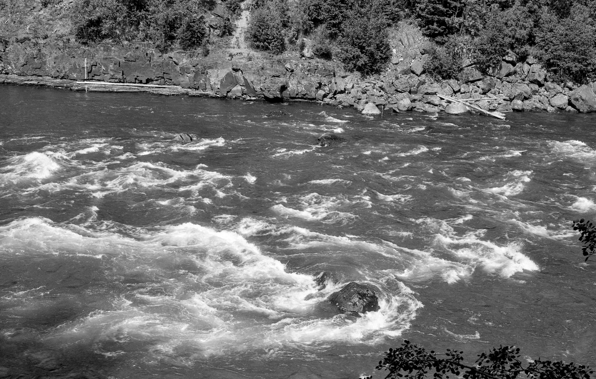 Elveparti fra Gudbrandsdalslågen nedenfor Hunderfossen.  Fotografiet er tatt fra en bratt elveskrent på østsida av vassdraget (Øyer-sida) mot et steinete elveløp der vannet passerte i stride strømmer med skumtopper.  Ved motsatt elvebredd - på Fåberg-sida - ser vi en steil bergvegg, og inntil den var det reist en loddrett målestav, antakelig for å registrere vekslinger i vannstanden.  Bildet ble tatt i 1961, antakelig med sikte på å dokumentere vannføringa i denne delen av Lågen før det prosjekterte kraftverket ved Hunderfossen ble ferdig et par år seinere.