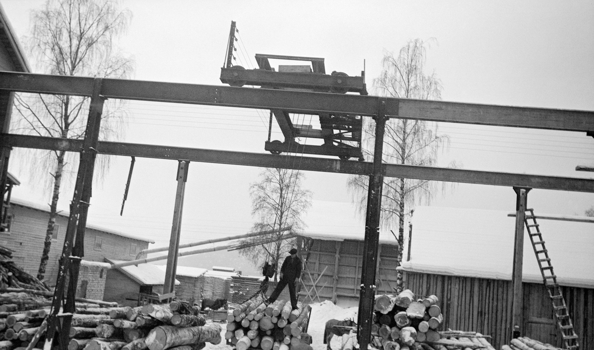 Kranbane ved foreløpig uidentifisert norsk sagbruk.  Kranbanen besto av to parallelle skinner, plassert på høye søyler av stål eller tre, på tvers av vegen inn mot sagbruket.  Oppå disse skinnene ser vi ei vogn av stål, og i denne vogna hang det ei kran med en løftekrok.  Da dette fotografiet ble tatt hadde en mann klatret opp på et tømmerlass og festet denne kroken i kjettinger som var slått rundt et lastebillass med tømmer.  Tømmeret skulle løftes og trilles inn på sagbruket, der stokkene skulle skjæres til trelast.  Vi ser saghuset til venstre i bildet, med rør over til flishuset sentralt i bildet.  Mellom disse to bygningene skimter vi noen av bordstablene på bruket.