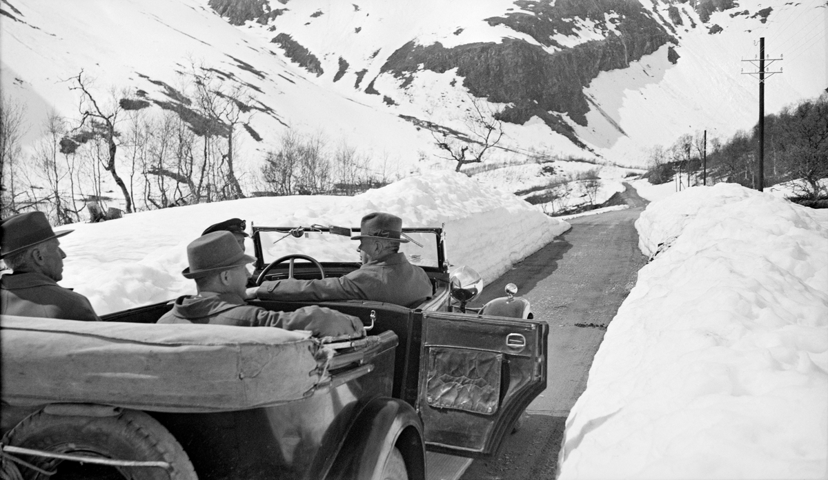 Utferd med fire menn, antakelig ansatte i administrasjonen eller styremedlemmer i Det norske Skogselskap, som i 1938 avviklet årsmøte på Sunnmøre med Møre skogselskap som vertskap.  Fotografiet er sannsynligvis tatt på en fjellovergang mens sentrale møtedeltakere var på veg mot Sunnmøre.  Vi ser fire menn i en åpen bil, som da dette fotografiet ble tatt sto parkert med den ene bakdøra åpen på en bar grusveg som var omgitt av høye snøplogkanter sjøl om bildet ble tatt seint på våren.