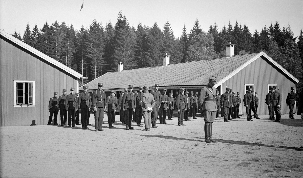 Mønstring av oppstilte soldater foran kaserner ved Ulven i Os i Hordaland.  Fotografiet er tatt under den såkalte skogrekruttskolen som ble avviklet her sommeren 1932.  Dette tiltaket samlet 86 rekrutter til ei blanding av militære øvelser og skogreisingsaktiviteter på statens eiendommer i området samt noen privateiendommer.  Skogrekruttskolen var lagt til et område der det lenge hadde vært arbeidet med skogreising.  Bak soldatene og kasernene ser vi en tettsluttet, snart hogstmoden granskog, som antakelig må ha tjent som et inspirerende eksempel på hvordan man kunne skape økonomiske verdier på ellers lavproduktive utmarksarealer.  Ulven ble underlagt den statlige skogforvaltninga på Vestlandet i 1872, og planting på denne kystnære lokaliteten hadde pågått siden den tid.  Fra samme år hadde fortsetaten latt Bergenske Brigade leie et areal på Ulven som ekserserplass mot en godtgjørelse på 400 spesidaler i året.  Kombinasjonen av skogreisings- og militæraktiviteter på dette stedet var med andre ord gammel da dette fotografiet ble tatt. Litt generell informasjon om skogrekruttskolen på Ulven i 1932 finnes under fanen «Opplysninger».