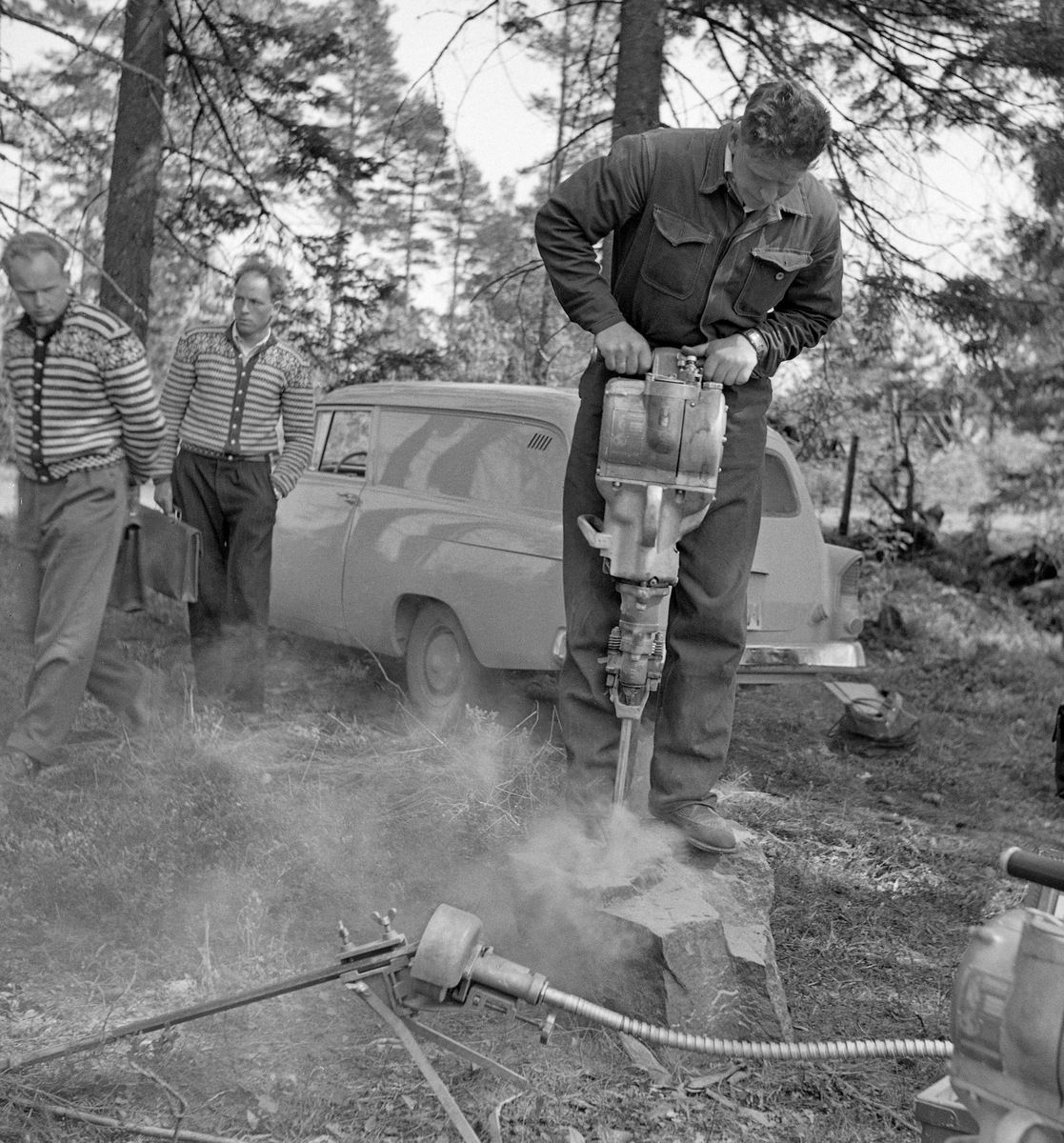 Fra demonstrasjon av bærbar bergbormskin for aktører fra skogbruket, antakelig tidlig i 19690-åra.  Fotografiet viser en kjeledresskledd mann som sto på en steinblokk med en maskin som han boret et hull med.  Finforstøvet stein sto som ei sky omkring føttene hans.  Ved inngangen til 1960-åra var det tre slike håndholdte bormaskintyper på det norske markedet: Engelske Warsop og svenske Pionjär og Atlas Copco.  Det vi ser på dette bildet er sannsynligvis en modell fra den sistnevnte produsenten, Atlas Copco Cobra.  Denne leverandøren hadde den letteste håndholdte bergbormaskinen på denne tida - cirka 24 kilo.  Den cirka 60 centimeter lange bormaskinen kunne lage hull som var inntil 4 meter djupe.  Bergboring var aktuelt i skogbruket i en periode da det ble sprengt en del digre steinblokker i forbindelse med bygging av skogsbilveger.  I bakgrunnen på dette bildet ser vi to karer med strikkejakker i såkalt Fana-mønster på overkroppen og med kontorvesker i hendene.  De fulgte boringa på respektfull avstand.  Vi ser også (skrått bakfra) også en Opel olympia (varebilvdersjonen av bilmodellen Opel rekord), antakelig en modell fra perioden 1958-1960.