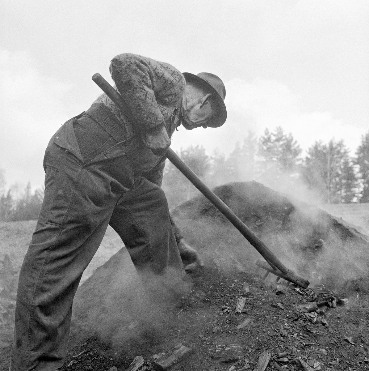 Trekolbrenning hos smeden Jørgen eller Jørn Romundstad (1890-1972) på Vegårshei i Aust-Agder.  Bildet skal være tatt våren 1968.  Smeden og kolbrenneren er fotografert i en fase da mila skulle rives.  Romundstad rakte vekk masse fra det rykende miledekket med ei langtinnet rive.  Ved sida av ham sto et jernspett og ei rive med noe bredere hode.  Romundstad var kledd i bomullsoverall og en genser med rombeformet mønster.  På hodet bar han en hatt.  Ei kort orientering om kolbrenning i slike miler finnes under fanen «Opplysninger».  Kullmile. Mile. Kolmile.