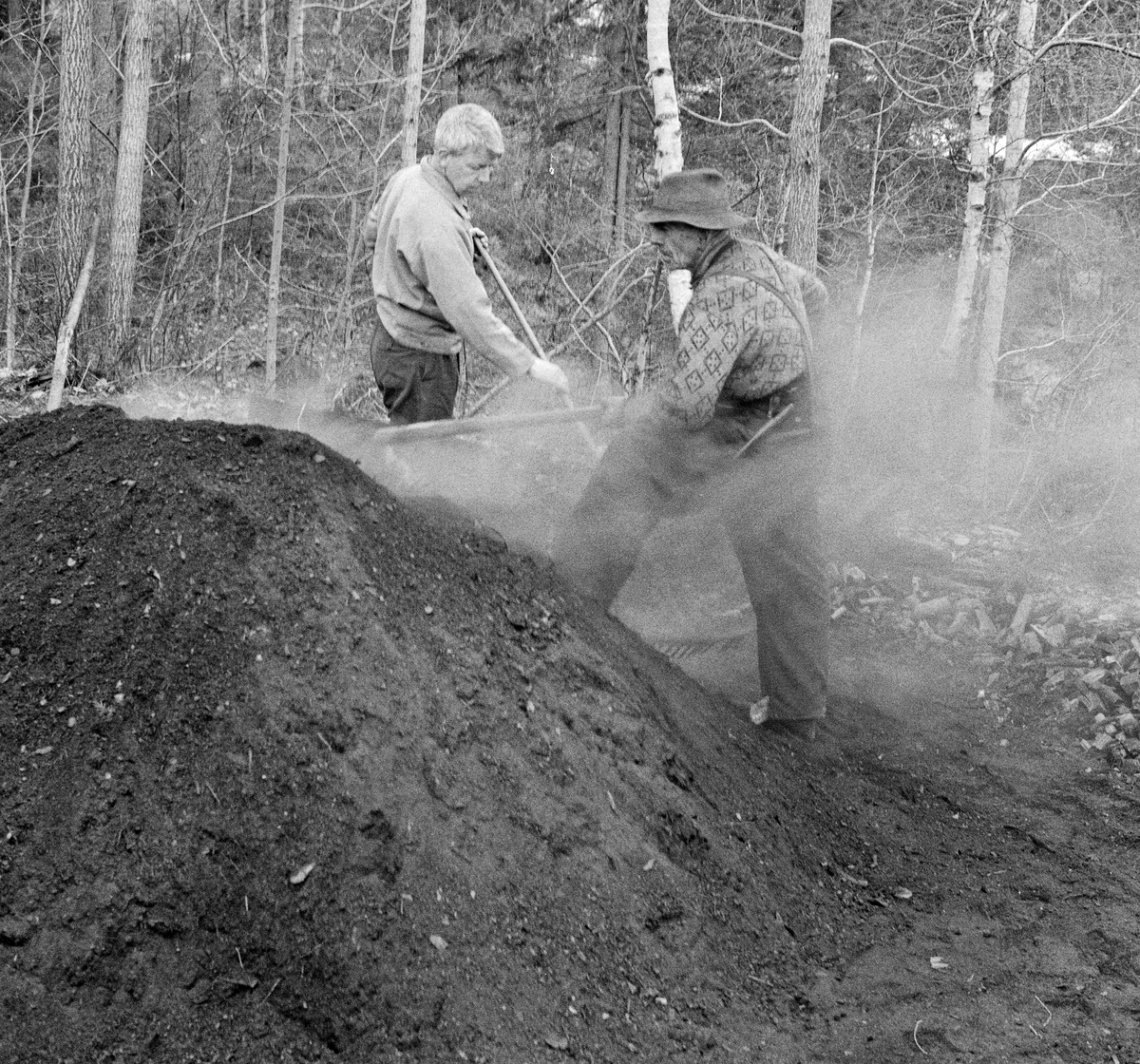 Trekolbrenning hos smeden Jørgen eller Jørn Romundstad (1890-1972) på Vegårshei i Aust-Agder.  Bildet skal være tatt våren 1968.  Vi ser smeden (til høyre) sammen med forstkandidaten og journalisten Andreas Vevstad (1930-2007), som var med museumsbestyrer Tore Fossum som kjentmann da han besøkte Romundstad.  Vevstad og Romundstad rakte kol på milebotnen med langtinnete river.  Begge karene var arbeidskledde.  Kullmile. Mile. Kolmile.