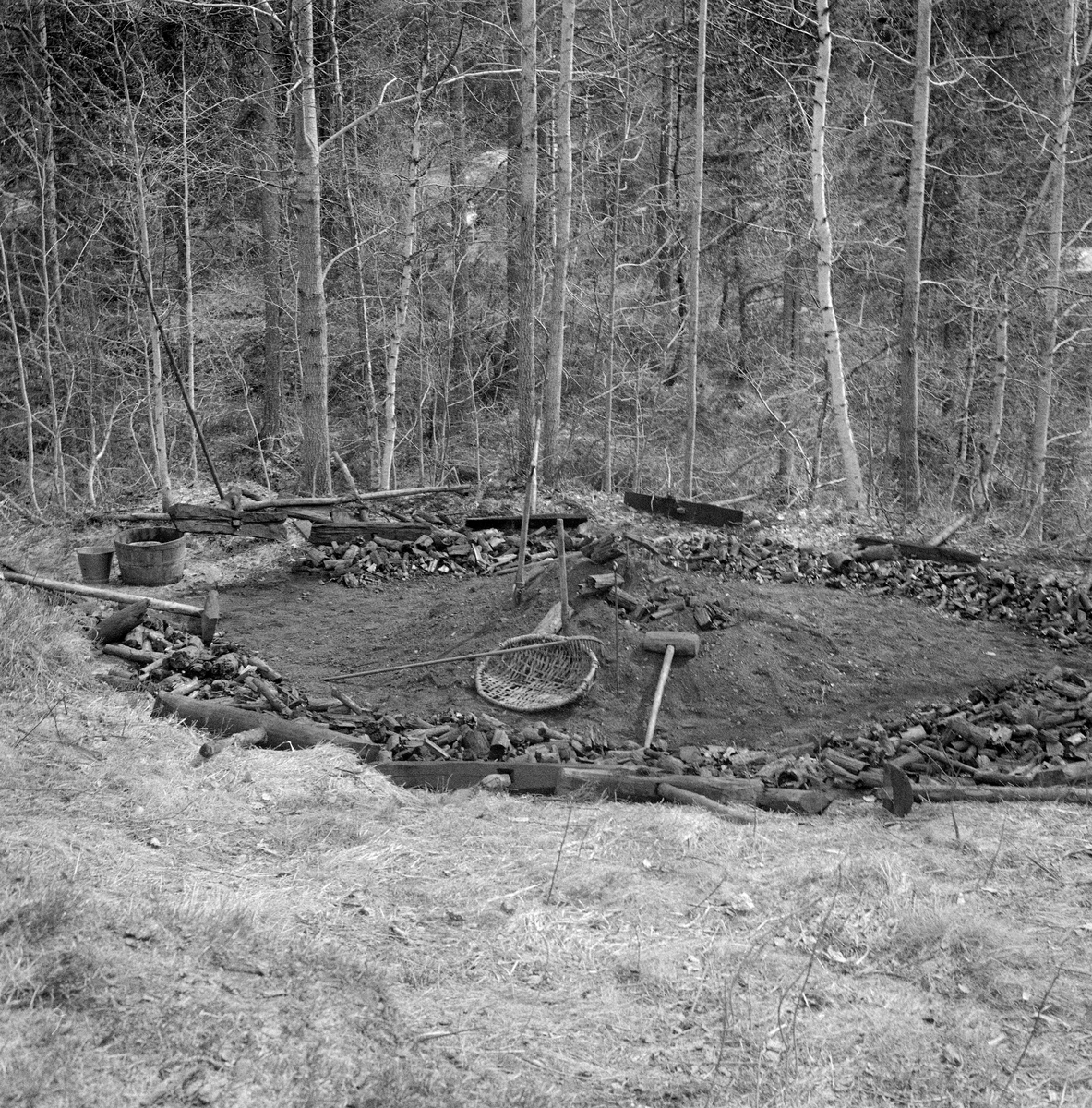 Kolbotn hos smeden Jørgen eller Jørn Romundstad (1890-1972) på Vegårshei i Aust-Agder.  Bildet skal være tatt våren 1968 mot den sirkelformete kolbotnen, der Romundstad hadde rakt kolbitene ut mot sidene.  På opptakstidspunktet lå det bare noen få kolbiter igjen i sentrum.  Framfor lå redskapene som ble brukt under kolbrenninga.  Vi ser et flettet kolfat, et greip, en spade, et spett, ei rive og ei treklubbe.  Vi ser også ei sinkbøtte og ei avskjæring av tre som inneholdt vann, for slokking dersom det skulle blusse opp varme i kolbitene.  I ytterkanten av kolbotnen lå det «stubbehyller», T-formete treelementer som skulle hindre at dekket på de bratte sidekantene av mila raste ned. Ei kort orientering om kolbrenning i slike miler finnes under fanen «Opplysninger». Trekolbrenning. Kullmile. Mile. Kolmile.