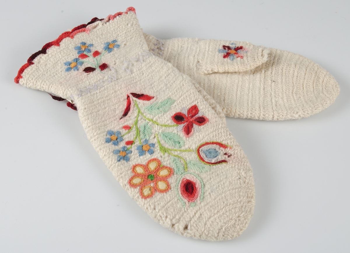 Ett par tumvantar i nålbindningsteknik tillverkade av vit ull och dekorerade med broderi i ullgarn. Den övre kanten har en virkad uddkant med rött ullgarn.