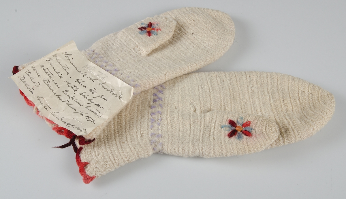 Ett par tumvantar i nålbindningsteknik tillverkade av vit ull och dekorerade med broderi i ullgarn. Den övre kanten har en virkad uddkant med rött ullgarn.