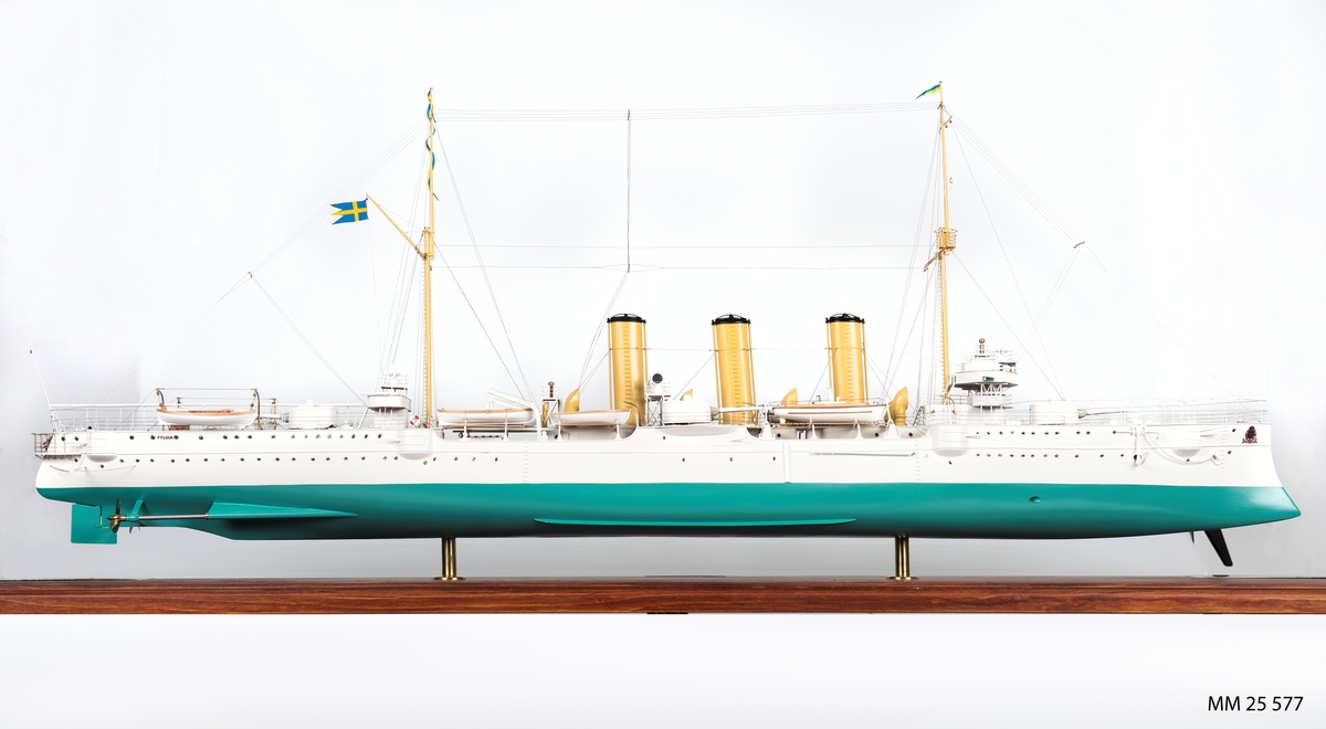 Fartygsmodell av pansarkryssaren Fylgia i skala 1:100. Detaljerad modell av Fylgia i originalutförandet. Vitt skrov, grön botten, tegelrött däck. Tre skorstenar samt master i gult. Tio skeppsbåtar, tio kassematter, kanoner, förstävsornament samt örlogsflagga och vimplar hissade. Modellen i monter av plexiglas samt botten av trä. Tre mässingsbrickor: 1. "Modellen byggd och planerad av Viktor von Sabsay - Harry Berlin". 2. "Pansarkryssaren Fylgia - Byggd av Finnboda varv, levererad 2 juni 1907, utrangerad 30 januari 1953. Deplacement: 4310/4980 ton. Dimensioner: 115,1/117.3 m x 148,8 m x 5,1 m. Maskineri och pannor: 2x4-CYL. Bergsunds stående trippelexp. kolvångmaskiner 12 000 IHK, 12 x Yarrow-pannor, 2 x Prop. Auktionsradie/fart: 8000nm/ 10 knop. Toppfart: 21,5 knop. Pansar: sida 100 mm. Kanontorn: 125 mm, Manövertorn: 100 mm. Däck: 35 mm. Bestyckning: 8-15,2 kan m/03o dubbeltorn. 14-57 mm kan m/89 B. 2-37 mm kan m/98 B. 4-45,7 cm torpedtuber m/04. Besättning: 322 man. Skala 1:100.". 3. "Befälhavare på första och sista resan".