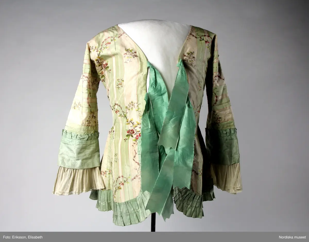 Katalogkort:
Kofta 1780-talet för fru J.Keyser f. Halling 1751-1797, gift 1770. Av bredrandig vit och ljusgrön taft med smala röda linjer samt glesa, slingrande blomrankor i färger. Helt fodrad med ljusrött glättat linnetyg. Koftan består endast av två stycken ( halva ryggen, ärmen och motsvarande stycke i ett, utan axelsöm, endast ryggsöm och sidsöm som övergår i ärmens innersöm.) hopknuten med rosetter kant i kant mitt fram med tre par breda ljusgröna sidenband. Kanten nedtill rundskuren med inlagda veck i rygg och sidor samt kilar i framstyckena. Ett veckat rysch av ljusgrön taft är påsytt runt kanten nedtill från den nedersta rosetten. Ärmen går över armbågen med bred manschett av ljusgrön taft med smalt veckat rysch i överkanten. Rynkade enkla engageanter av glest, vitt, randat linnetyg med smal knypplad spets i kanten. Obetydligt lagad å ärmarna. Byst 90 cm, midja 80 cm. Tygets vådbredd minst 56 cm, tygåtgång vid denna bredd 2,55 m.

