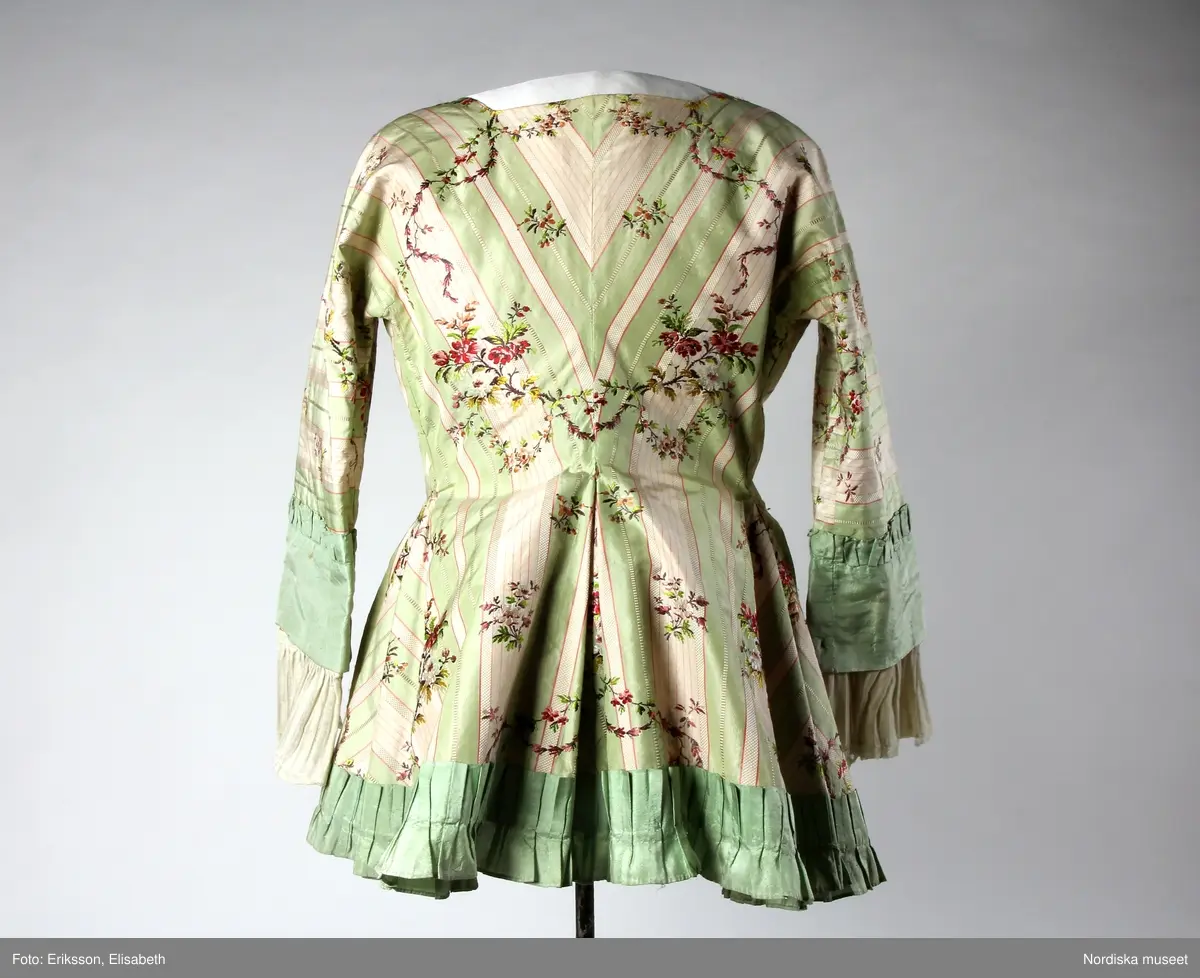 Katalogkort:
Kofta 1780-talet för fru J.Keyser f. Halling 1751-1797, gift 1770. Av bredrandig vit och ljusgrön taft med smala röda linjer samt glesa, slingrande blomrankor i färger. Helt fodrad med ljusrött glättat linnetyg. Koftan består endast av två stycken ( halva ryggen, ärmen och motsvarande stycke i ett, utan axelsöm, endast ryggsöm och sidsöm som övergår i ärmens innersöm.) hopknuten med rosetter kant i kant mitt fram med tre par breda ljusgröna sidenband. Kanten nedtill rundskuren med inlagda veck i rygg och sidor samt kilar i framstyckena. Ett veckat rysch av ljusgrön taft är påsytt runt kanten nedtill från den nedersta rosetten. Ärmen går över armbågen med bred manschett av ljusgrön taft med smalt veckat rysch i överkanten. Rynkade enkla engageanter av glest, vitt, randat linnetyg med smal knypplad spets i kanten. Obetydligt lagad å ärmarna. Byst 90 cm, midja 80 cm. Tygets vådbredd minst 56 cm, tygåtgång vid denna bredd 2,55 m.

