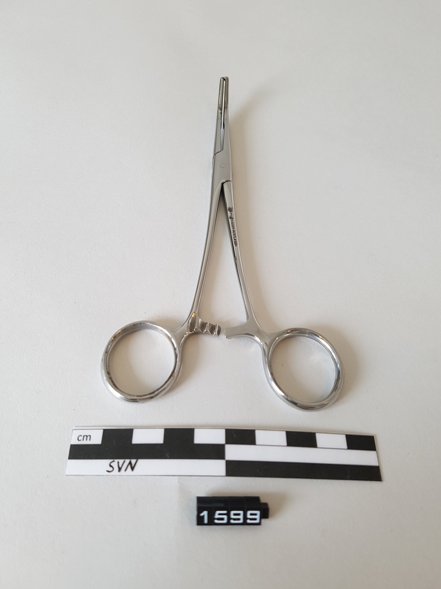 Gjenstanden er en kirurgisk bøyd klemme. 
Klemmen er utformet som en saks, men i stede for skjærekniver er det to flater som møtes og klemmes sammen.
Flatene som møtes har serrateringer, riller. ytterst på klemmen har flatene tenner som går i hverandre.
To tenner på en side og en tann på den andre siden.
Gjenstanden har fingerhull for manipulering av objektet.
like over fingerhullene er det en låsemekanisme med en hake på den ene delen og flere motstående haker på den andre delen
Instrumentet kan låses ved hjelp av disse og hvor mye press det legges på tangspissen bestemmes av hvilken av mothakene 
det låses mot.